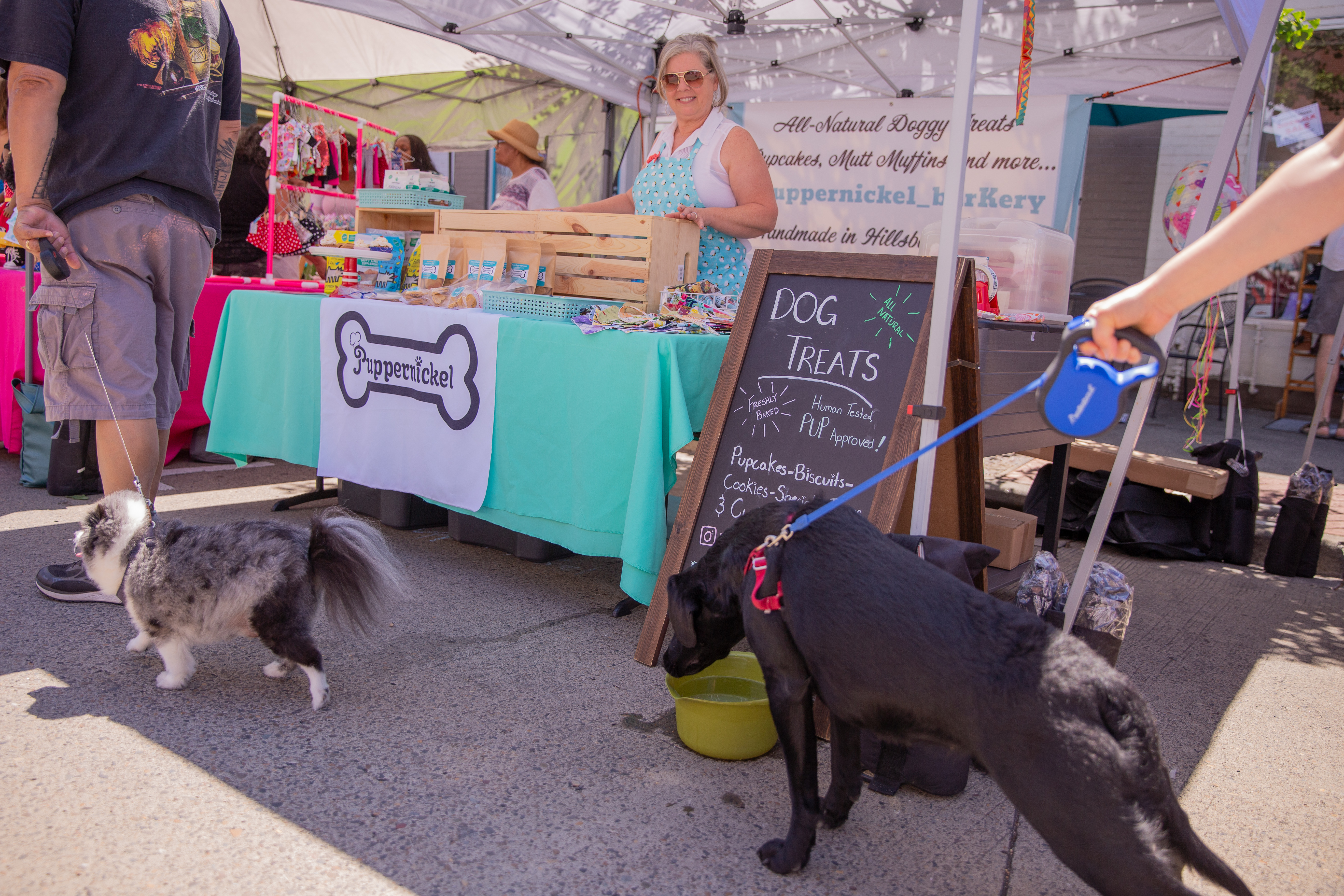 Krystal, the owner of Pumpernickel, stands in her booth full of treats for dogs at the Farmers Market, smiling at a black dog stretching it's neck to reach the bowl of water she's provided. 