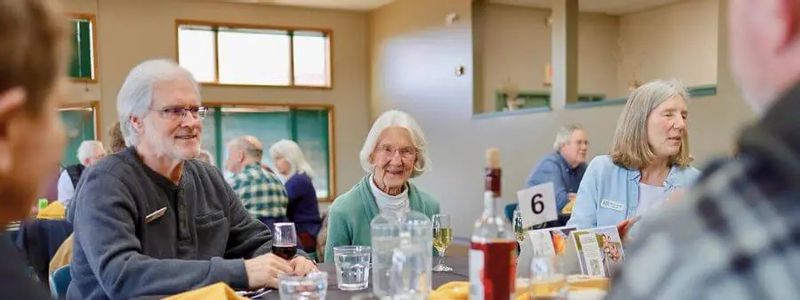 A group of older adults sit around a round table in a dining hall, chatting and enjoying drinks during a winter group event, with place settings, glasses, and bottles on the table.