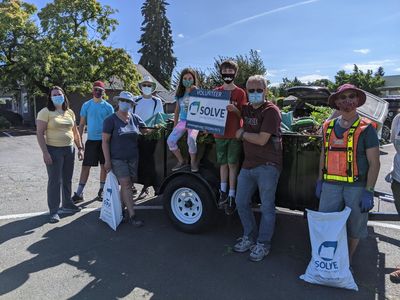 Eight volunteers of various ages standing next to a black utility trailer filled with yard debris. One child is holding a SOLVE Oregon sign and two of the adults have SOLVE Oregon bags. SOLVE garbage 