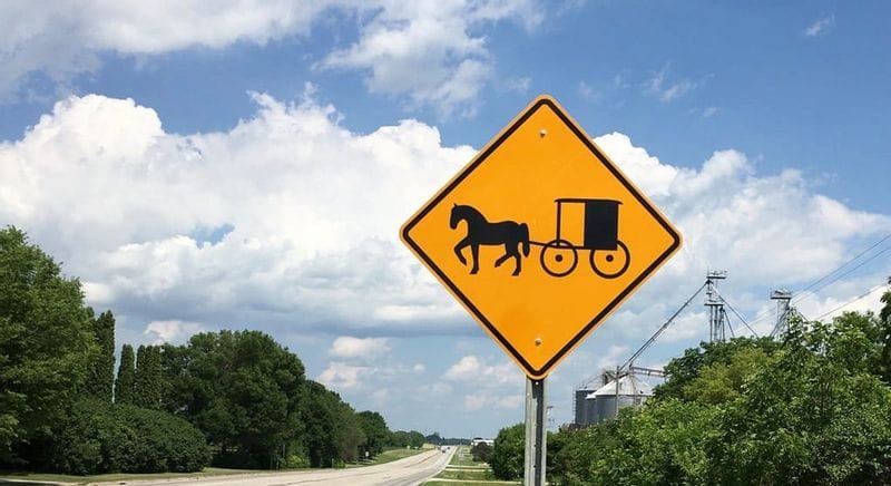 Amish buggy crossing warning sign beside a rural highway, showing a horse pulling a traditional Amish carriage silhouette on a yellow diamond traffic sign, with green roadside grass, trees, grain silos in the distance, and a partly cloudy blue sky.