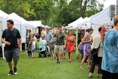 Crowds stroll through Art in the Park in Lanesboro, browsing white-tent artist booths under leafy trees, with families, children, and a stroller moving along the grassy park pathway during the outdoor arts festival.