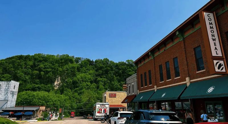 The Commonweal Theatre building lines downtown Lanesboro’s main street, with brick storefronts, parked cars, and a wooded bluff rising behind the town on a clear summer day.