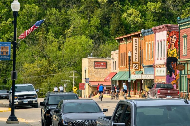 Cyclists ride through downtown Lanesboro past the Commonweal Theatre, Stone Mill Hotel & Suites, colorful storefronts, and spring-green bluffs.