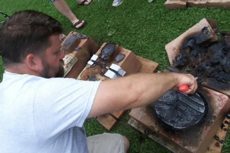 A man demonstrates metal casting during the Sculpture Medallion Tour in Lanesboro, pouring molten material into a mold set on bricks outdoors.