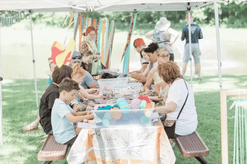 Families and children gather under a canopy creating art together during Art in the Park in Lanesboro, seated at picnic tables with craft supplies and colorful materials.
