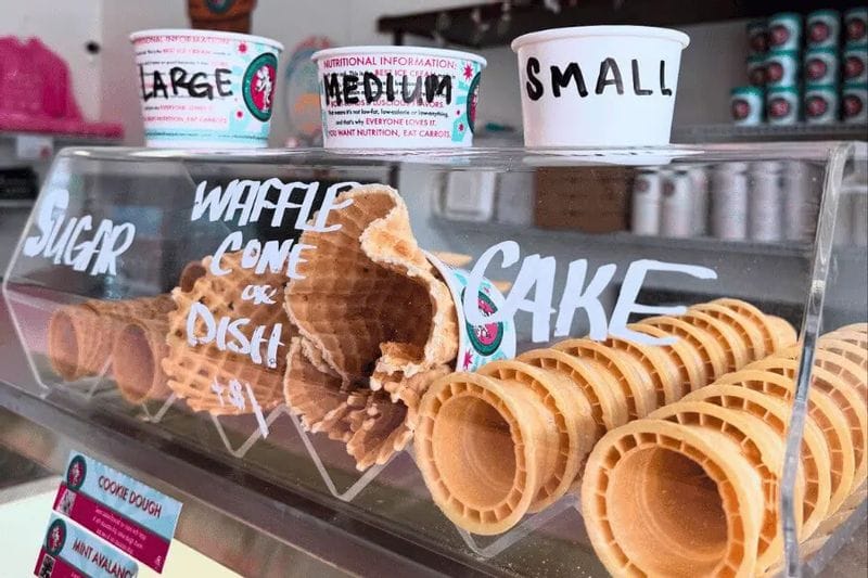 Display case of waffle cones and cake cones at Clara’s Eatery in Lanesboro, Minnesota, with hand-lettered signage and ice cream cup size labels visible above the cones.