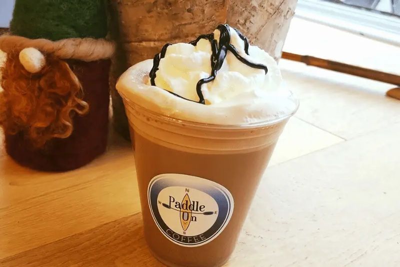 Iced coffee topped with whipped cream and chocolate drizzle in a clear cup from Paddle On Coffee, sitting on a wooden counter inside the café in Lanesboro, Minnesota.