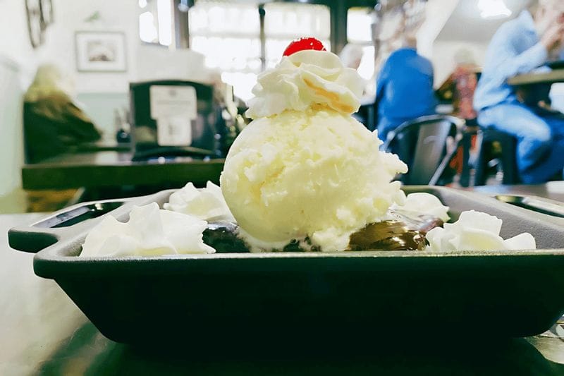 Warm dessert topped with a scoop of vanilla ice cream, whipped cream, and a cherry, served on a black plate inside Pedal Pushers Café in Lanesboro, Minnesota.