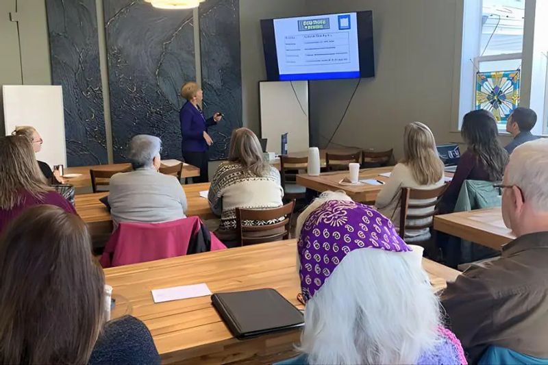 Group of adults seated at wooden tables in a café meeting room while a presenter points to a slideshow on a wall-mounted screen during a workshop or community presentation.