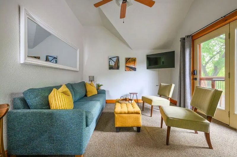 Bright, modern hotel suite with a teal sofa, yellow accent pillows and ottoman, two lounge chairs, wall-mounted TV, ceiling fan, and glass door opening to a private balcony with trees outside.