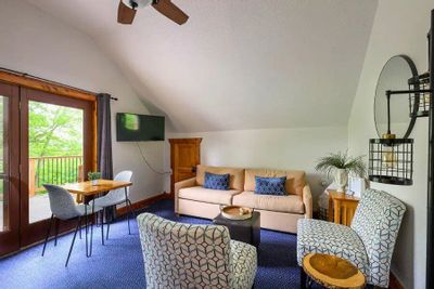 Bright hotel suite with a seating area featuring a sofa, patterned armchairs, small dining table, and a wall-mounted TV, with glass doors opening to a balcony overlooking trees.