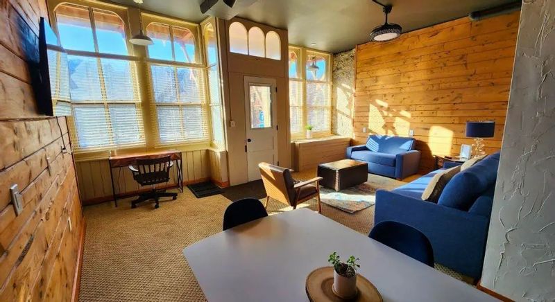Sunlit living area with wood-paneled walls, large windows, a private patio door, blue sofas, a small dining table, and a desk, creating a cozy, modern suite interior.