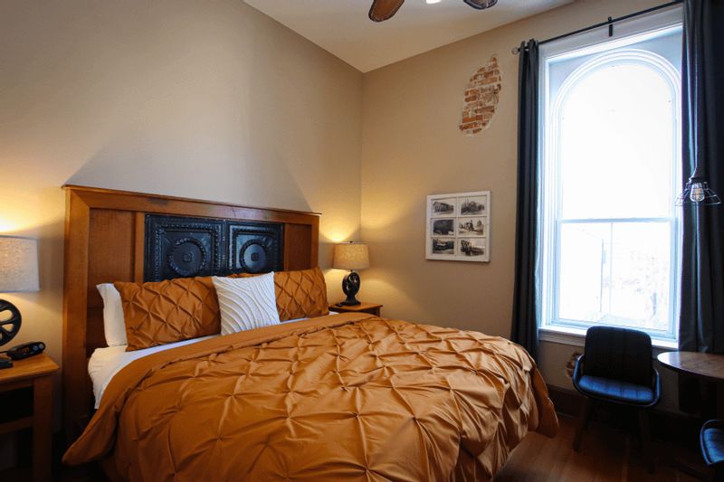 The Iron Horse King Room at the Root River Inn features a cozy bedroom with a king bed, warm-toned bedding, decorative headboard, tall arched window, and vintage-inspired accents that reflect the building’s historic character.