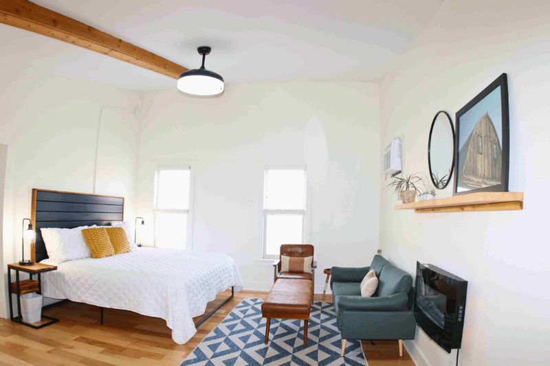 The Mayoral Suite at the Root River Inn features a bright, modern bedroom with a comfortable bed, sitting area, wood floors, exposed beam detail, and large windows that fill the space with natural light.