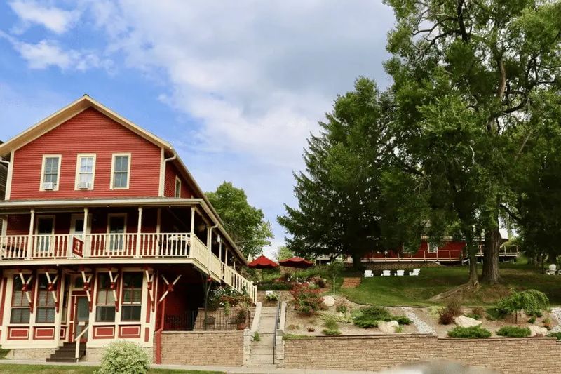 A street-level view of the Root River Inn shows its historic red building with a wraparound porch, landscaped hillside, and outdoor seating areas set among mature trees in Lanesboro.