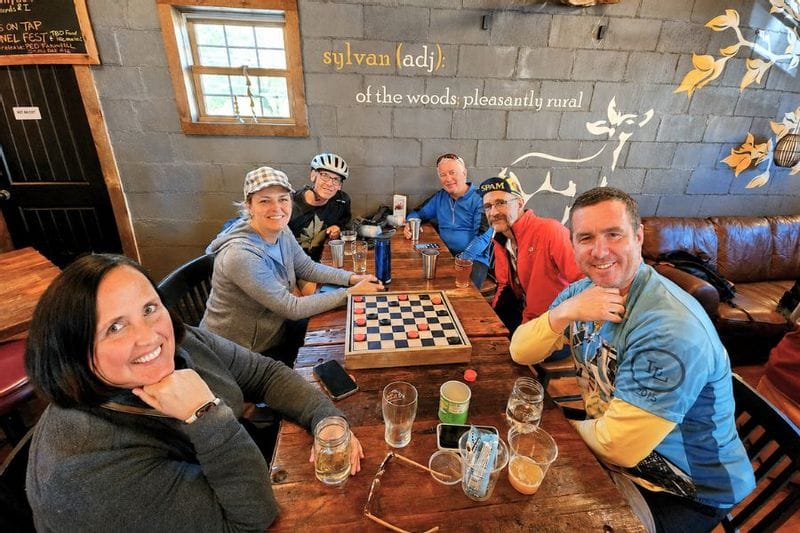 A group of people inside Sylvan Brewing enjoying beer and playing checkers. Some have bike helmets and great on, all are looking at the camera smiling.