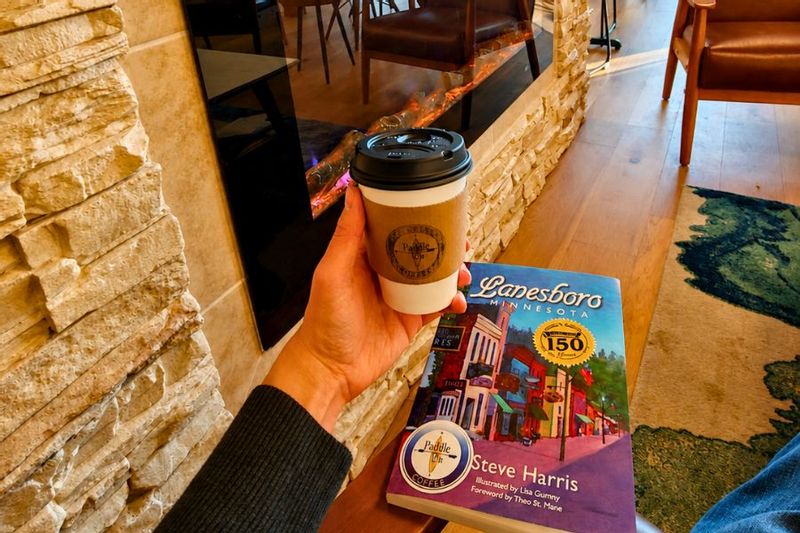 Hand holding a coffee near a fireplace with the book Lanesboro, Minnesota by Steve Harris on a table in a cozy indoor setting.