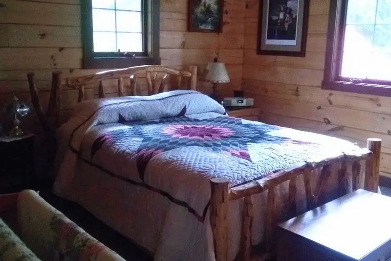 Rustic bedroom inside a log cabin featuring a handcrafted wooden bed, quilted bedspread, wood-paneled walls, and soft natural light from nearby windows.