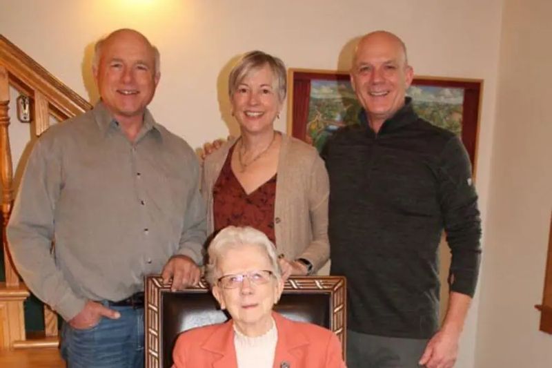 Members of the Bunge family pose together indoors at the Cottage House Inn, with three adults standing behind an elderly woman seated in a chair, smiling in a warmly lit room.