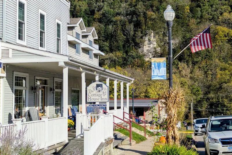 Exterior of the Cottage House Inn in Lanesboro during fall, with a white porch, inn signage, autumn decorations, an American flag, and wooded bluffs in the background.