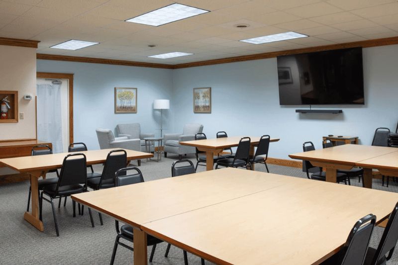 Meeting room at the Cottage House Inn with rectangular tables, black chairs, a wall-mounted TV, and a small seating area with armchairs and framed artwork.