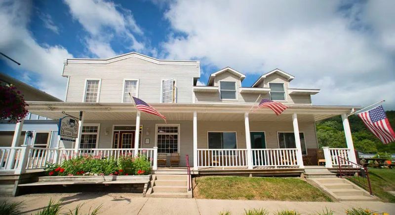 Exterior of the Cottage House Inn in Lanesboro, Minnesota, featuring a white historic inn with a wraparound porch, American flags, flower beds, and a sunny blue sky overhead.