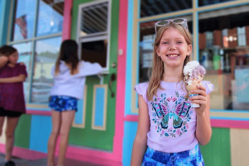 A smiling child holds a cone of ice cream outside Another Time Ice Cream Parlor in downtown Lanesboro, with the shop’s colorful pink, teal, and green storefront in the background on a summer day.
