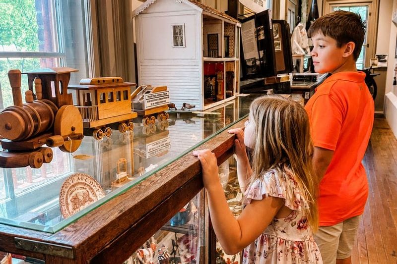 Two children inside the Lanesboro History  Museum looking at a historical display featuring a model wooden train on top of a glass case containing other historical displays