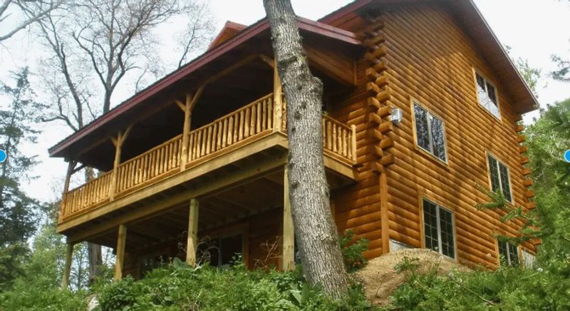 Log cabin with a wraparound porch perched on a wooded hillside in the Root River Valley, surrounded by mature trees and lush greenery near Lanesboro, Minnesota.