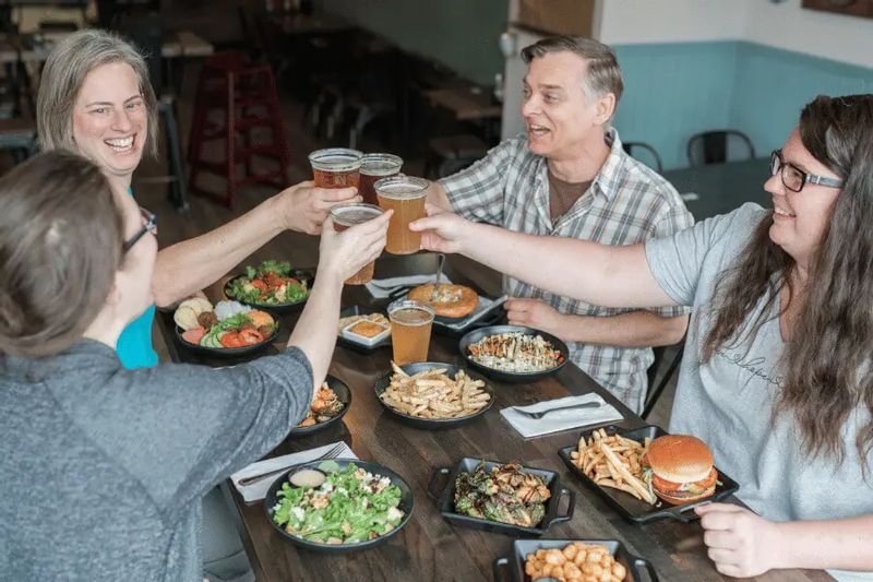 A group of friends raise glasses of beer in a toast around a table at Pedal Pushers Café, surrounded by burgers, fries, salads, and shared plates in a relaxed Lanesboro restaurant setting.