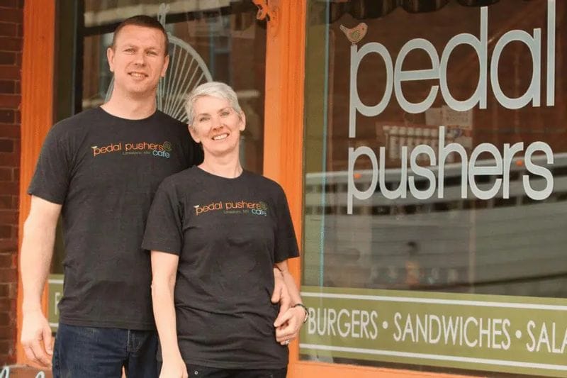Two owners stand smiling outside Pedal Pushers Café, wearing café-branded shirts in front of the restaurant’s window signage in downtown Lanesboro, Minnesota.