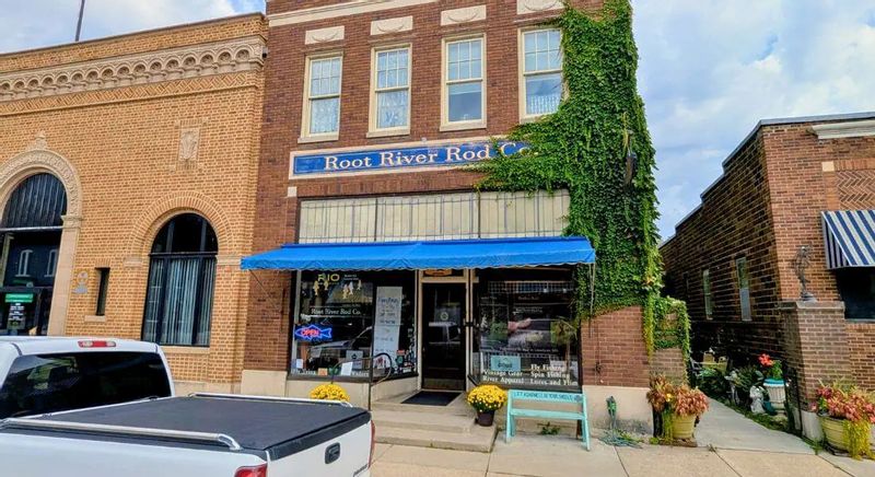 The storefront of Root River Rod Co. in downtown Lanesboro, featuring a brick building with a blue awning, ivy climbing the facade, fishing gear displays in the windows, and flower planters along the sidewalk.