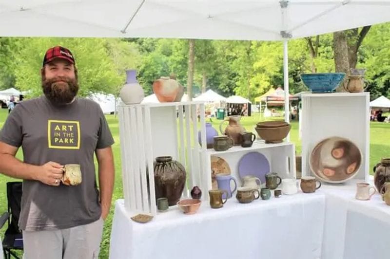 An artist stands beside a booth displaying handmade pottery and ceramic pieces at Art in the Park in Lanesboro, with white tents and green park space visible in the background.