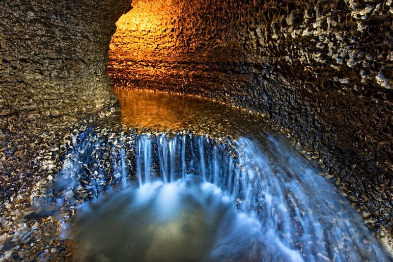 Underground waterfall flows over textured limestone inside Niagara Cave near Harmony, Minnesota, with warm cave lighting illuminating the rocky passage and pool below.