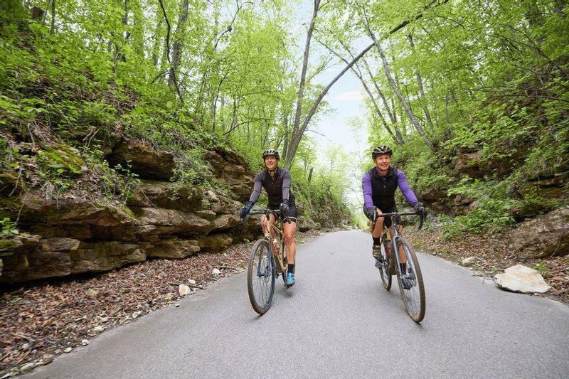 Two bikers riding on a paved trail through a wooden area with exposed rockfaces on either side