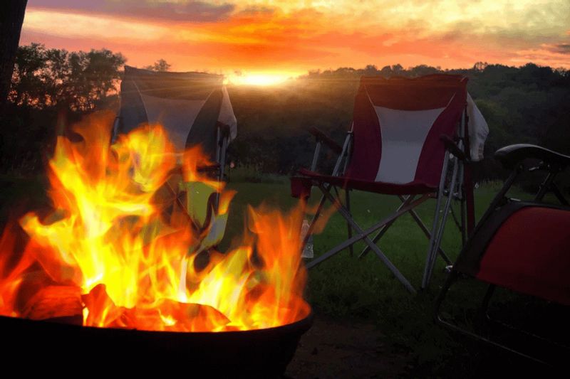 A glowing campfire burns at sunset beside folding camp chairs, creating a cozy evening scene at Eagle Cliff Campground with rolling hills and colorful skies in the background.