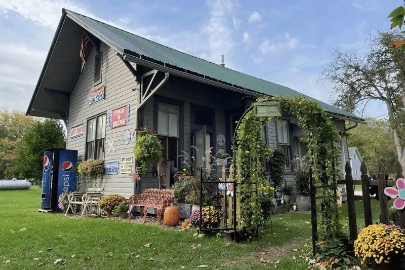 The Whalan Depot, a historic small-town building decorated with flowers, pumpkins, and vintage signs, sits along a grassy lawn in Whalan, Minnesota, welcoming visitors to shop and explore.