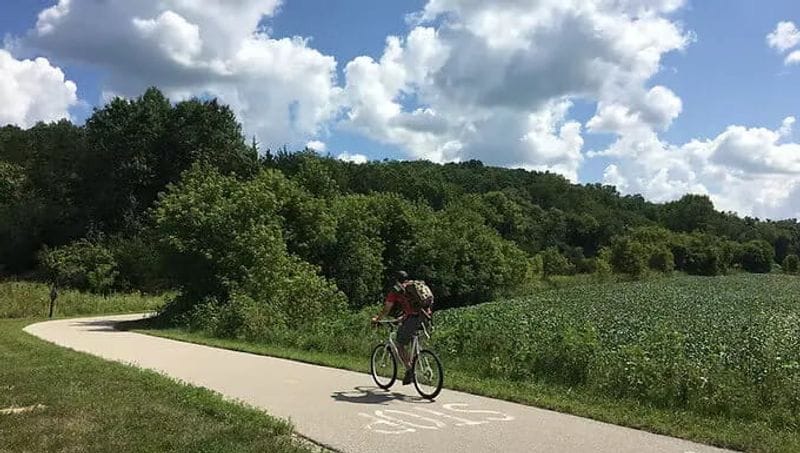 A cyclist rides along a paved trail through open countryside near Lanesboro, with rolling green hills, fields, and a partly cloudy summer sky in the background.