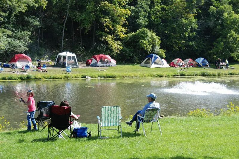 Families camp and relax along the water at Sylvan Park in Lanesboro, with tents set up on the grassy shore and children fishing beside the pond on a sunny day.