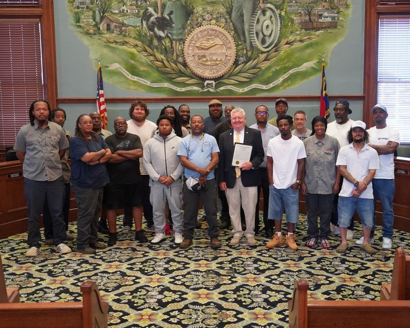 Sanitation workers stand with Mayor Doug Hendrix in the Statesville City Council Chambers during the June 15, 2026 City Council meeting. Mayor Hendrix holds a proclamation recognizing June 15–20, 2026, as National Waste and Recycling Workers Week, honoring the essential contributions of sanitation and recycling employees who help keep the community clean, safe, and healthy. The group is gathered beneath the City of Statesville seal, commemorating the recognition.