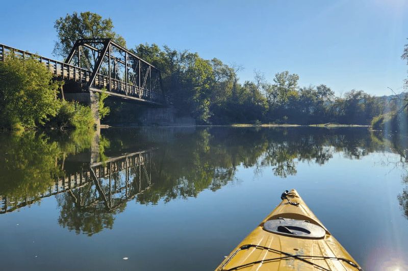 A kayak floats on the calm waters of the Root River near Lanesboro, with a steel truss bridge and tree-lined riverbanks reflected in the water on a clear day.