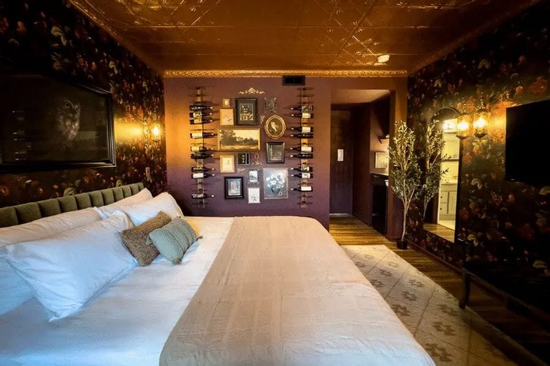 A stylish hotel bedroom features a large bed with layered pillows, dark floral wallpaper, warm ambient lighting, and a decorative wall displaying wine bottles and framed artwork, creating an intimate, upscale atmosphere.