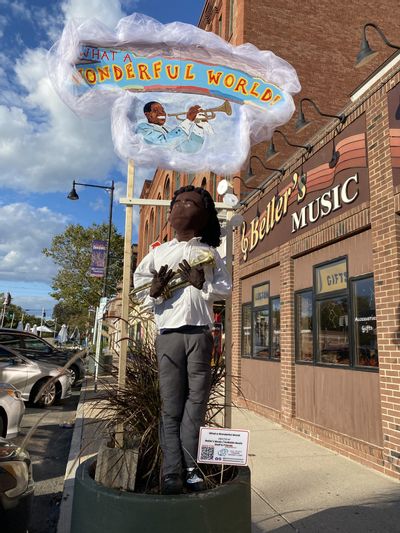 A sculpture is displayed in an outdoor planter. A sign reads What a wonderful world. The sculpture is a Black man holding a trumpet. In the background a storefront sign reads Beller's Music.