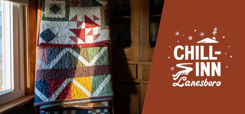 A colorful handmade quilt hangs by a window beside the Chill Inn Lanesboro logo, highlighting Lanesboro’s arts experience and cozy lodging atmosphere.