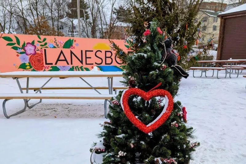 A decorated evergreen tree with a red heart ornament stands in the snow near the Lanesboro mural during winter, blending Valentine-themed décor with the town’s public art.
