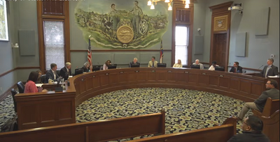 Members of the Statesville City Council sit around the curved council dais during a meeting inside City Hall. City staff, council members, and attendees are visible throughout the chamber beneath the large City of Statesville mural displayed on the wall behind the council seating area.