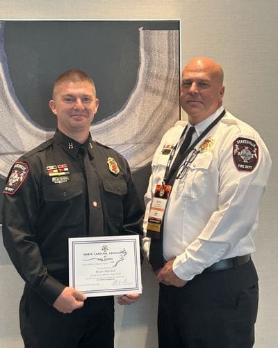 Captain B. Mitchell of the Statesville Fire Department stands beside Deputy Chief A. Kennedy while holding a certificate from the North Carolina Association of Fire Chiefs. The photo was taken during a formal recognition event honoring Captain Mitchell for earning his national fire officer credential. Both men are dressed in Statesville Fire Department uniforms and posed indoors in front of framed artwork.