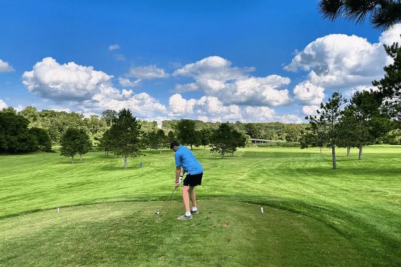 A golfer tees off on a wide, green golf course at the Old Barn Resort near Lanesboro, Minnesota, surrounded by rolling hills, trees, and a bright blue sky.