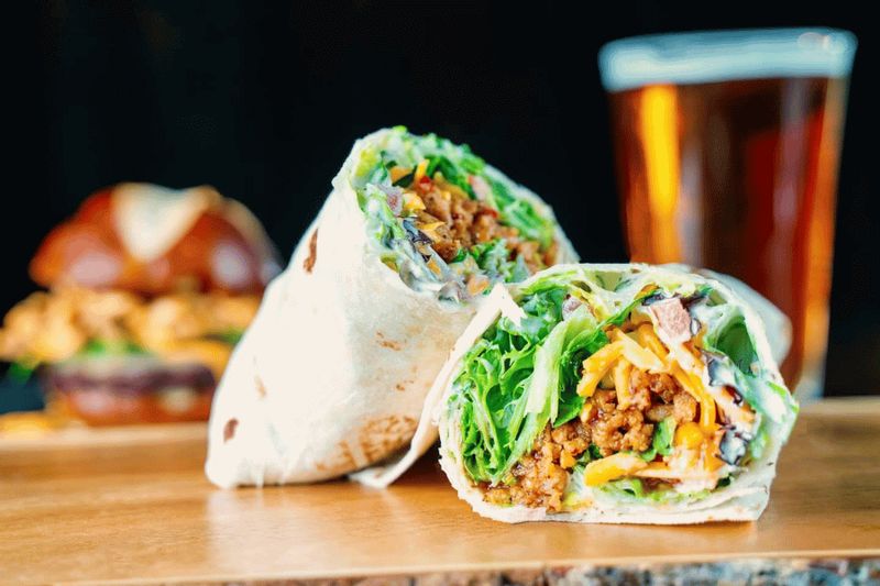 A fresh wrap filled with seasoned meat, lettuce, and shredded cheese is sliced in half on a wooden table, with a craft beer and burger softly blurred in the background at a local restaurant.