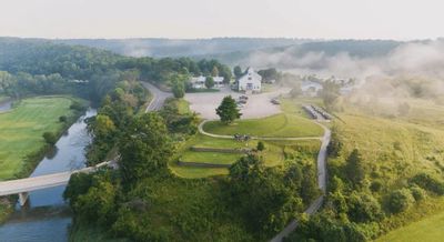 An aerial view of the Old Barn Resort near Lanesboro, Minnesota, shows a white barn-style venue surrounded by rolling hills, wooded bluffs, winding roads, and the Root River on a misty morning.